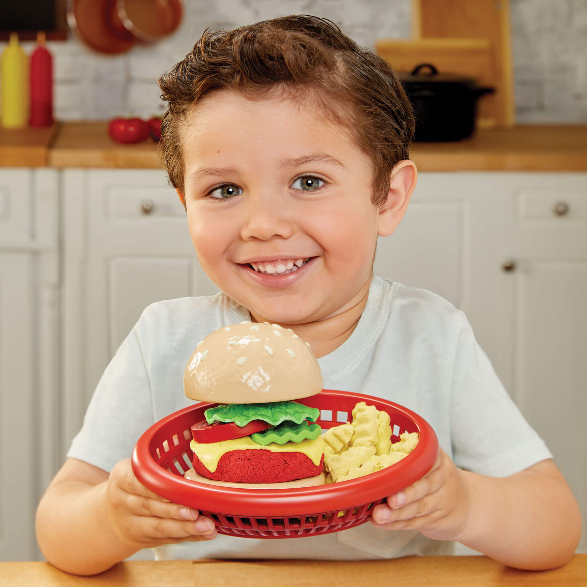 Child holding a toy hamburger set in a red basket in a kitchen setting