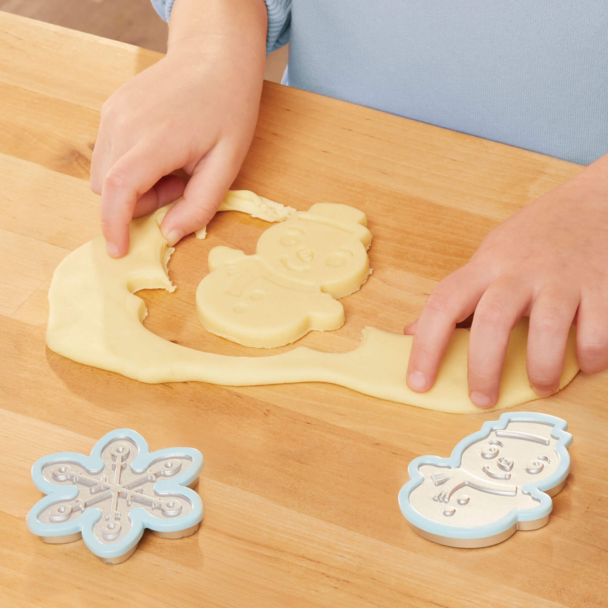 Hands shaping cookie dough with snowflake and snowman cookie cutters on a wooden table.